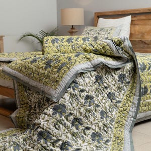 May include: A floral patterned quilt set in shades of yellow, gray, and white. The quilt features a repeating flower design and a striped border. The bedding is displayed on a wooden bed frame, suggesting a bedroom setting.