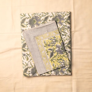 May include: A folded textile featuring a large floral pattern in shades of grey and yellow on a white background. The top layer displays a border with grey and white stripes, while the underlying fabric shows a repeating grey and yellow floral print.
