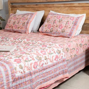 May include: A floral patterned quilt and two matching pillowcases in shades of pink, white, and green are displayed on a wooden bed frame. The bedding features a repeating flower design and a border.