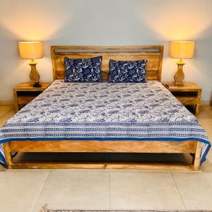 May include: A wooden bed frame with a headboard, featuring a white and blue floral patterned bedspread and two matching pillows. Two wooden nightstands are positioned on either side of the bed, each topped with a lamp emitting a warm glow.