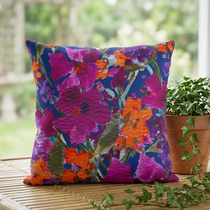 Puede incluir: Cojín decorativo cuadrado con un vibrante estampado floral. El cojín presenta grandes flores en tonos magenta, naranja y morado sobre un fondo azul oscuro. Se coloca sobre una mesa de bambú, con una planta en maceta cerca.