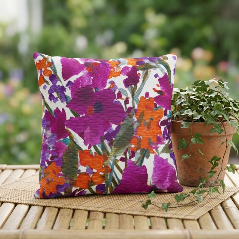 May include: Square decorative pillow with a vibrant floral pattern in shades of purple, orange, and green on a white background. The pillow is placed on a bamboo mat next to a potted plant.