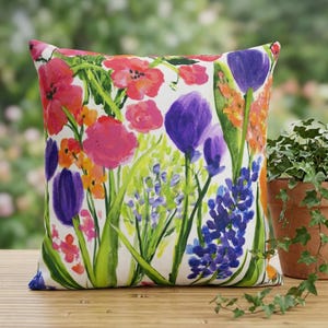 Pode incluir: Almofada decorativa quadrada com uma estampa floral vibrante. O design apresenta flores coloridas em tons de rosa, roxo, laranja e azul sobre um fundo branco. Folhas e caules verdes também são visíveis. A almofada está colocada sobre uma superfície de madeira.