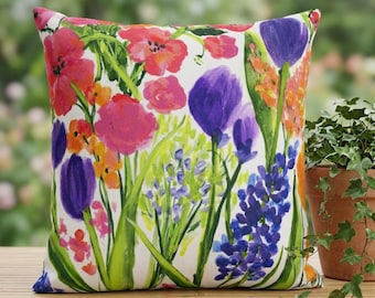 May include: Square decorative pillow with a vibrant floral print. The design features colorful flowers in shades of pink, purple, orange, and blue against a white background. Green leaves and stems are also visible. The pillow is placed on a wooden surface.