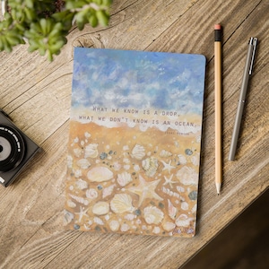 Puede incluir: Un cuaderno con un diseño pintado de una escena de playa con conchas marinas y una estrella de mar. La cita "Lo que sabemos es una gota, lo que no sabemos es un océano." de Isaac Newton está impresa en la portada.