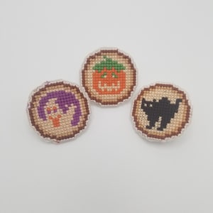 May include: Three small, round, embroidered patches with Halloween themes. The first patch features a purple ghost with orange eyes. The second patch features an orange pumpkin with a green stem and a jack-o-lantern face. The third patch features a black cat with white eyes.