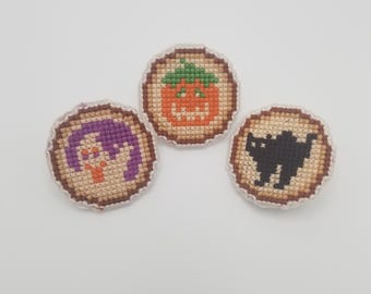 May include: Three small, round, embroidered patches with Halloween themes. The first patch features a purple ghost with orange eyes. The second patch features an orange pumpkin with a green stem and a jack-o-lantern face. The third patch features a black cat with white eyes.