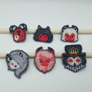 May include: Set of seven embroidered patches featuring cartoon characters with red, black, and white details. The characters have different expressions and are stitched on a gray fabric.