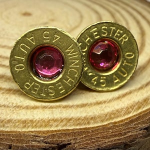 May include: A pair of brass-coloured stud earrings. Each earring is made from a .45 AUTO Winchester casing and features a round, pink gemstone in the centre. The earrings are displayed on a wooden surface.