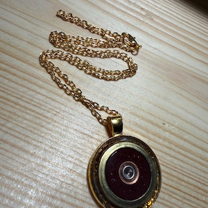 Könnte beinhalten: Eine goldfarbene Halskette mit einem runden Anhänger. Der Anhänger besteht aus dem Boden einer dunkelroten Schrotpatronenhülse mit einem goldfarbenen Metallrand und einem silberfarbenen Zündhütchen in der Mitte, an einer Gliederkette.