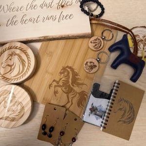 May include: A collection of horse-themed items, including wooden coasters, a cutting board, keychains, earrings, a leather horse figurine, and a notebook. The wooden sign reads "Where the dust flies & the heart runs free."
