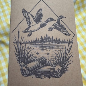 Puede incluir: Un cuaderno color canela con una ilustración en tinta negra de dos patos volando sobre un lago, un bosque y espadañas. Dos cartuchos de escopeta están colocados en primer plano en la parte inferior del diseño, todo dentro de un rombo.