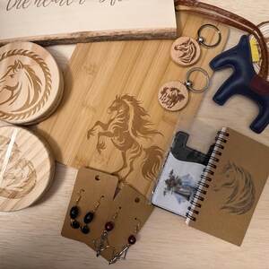 May include: A collection of horse-themed items, including wooden coasters, a cutting board, keychains, a leather horse figurine, earrings, a notebook, and a cardholder. The items feature engraved horse designs and are arranged on a light wooden surface.