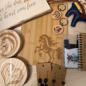 May include: A collection of horse-themed items, including a wooden cutting board with a horse engraving, keychains, coasters, earrings, a bracelet, and a small notebook. The cutting board has the text "the dust flies & the heart runs free".
