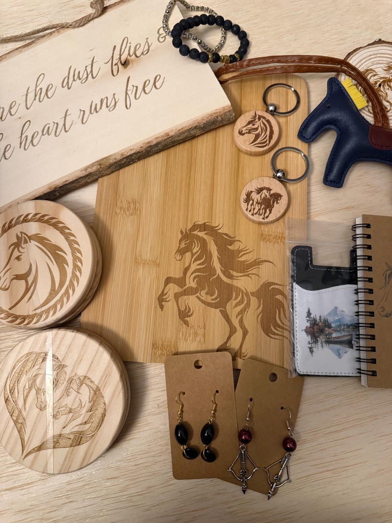 May include: A collection of horse-themed items, including a wooden cutting board with a horse engraving, keychains, coasters, earrings, a bracelet, and a small notebook. The cutting board has the text "the dust flies & the heart runs free".