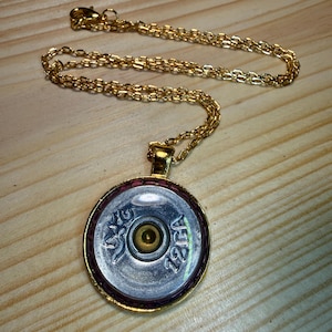 Könnte beinhalten: Eine goldfarbene Halskette mit einem runden Anhänger aus dem Boden einer 12-Gauge-Schrotpatrone. Die metallisch silberfarbene Mitte der Patronenhülse ist von einem goldfarbenen Rand umgeben und an einer passenden Gliederkette befestigt.
