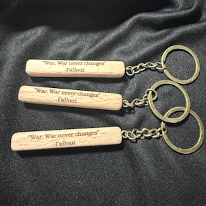 Könnte beinhalten: Drei Holzschlüsselanhänger mit dem eingravierten Text "War. War never changes" und "-Fallout". Jeder Schlüsselanhänger hat eine Metallkette und einen messingfarbenen Ring.