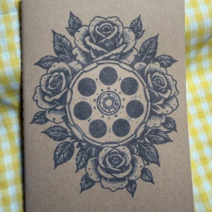 Puede incluir: Un cuaderno rectangular con cubierta de color tostado que presenta una ilustración en tinta negra de un tambor de revólver rodeado por un grupo de rosas en flor y hojas. El diseño está centrado en la portada con líneas detalladas.
