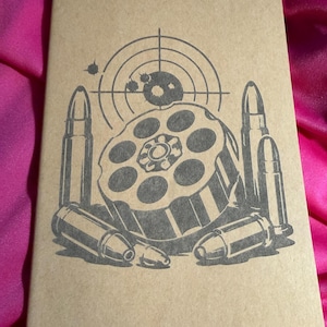 Puede incluir: Un cuaderno rectangular de color tostado con un estampado gráfico gris oscuro en la portada. La ilustración muestra un tambor de revólver rodeado de varias balas y una retícula de objetivo circular sobre el diseño del tambor.
