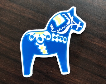 May include: A blue Dala horse sticker featuring white and yellow decorative patterns on the body, mane, and saddle area. The design includes stylized floral motifs and geometric lines, all outlined in a clean white border against a dark background.