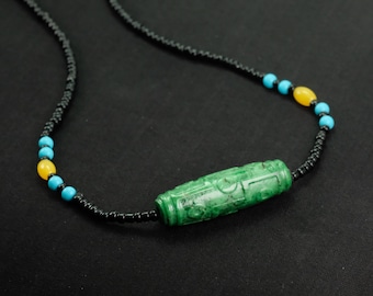 Puede incluir: Un collar con un colgante de jade verde tallado en forma cilíndrica. El collar presenta cuentas negras, intercaladas con cuentas turquesas y amarillas. El collar se muestra sobre una superficie negra.