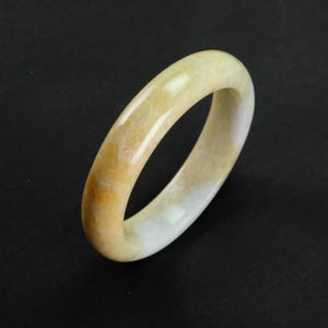 Op de afbeelding: Een gladde, gepolijste jade armband. De armband heeft een mix van crèmewitte en goudgele tinten. Het cirkelvormige ontwerp is eenvoudig en elegant, geschikt voor dagelijks gebruik. De jade is een natuursteen.