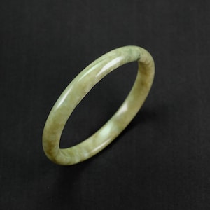 Könnte beinhalten: Ein glattes, hellgrünes Jade-Armband. Das Armband ist aus poliertem Jade gefertigt und hat ein subtiles, wirbelndes Muster.
