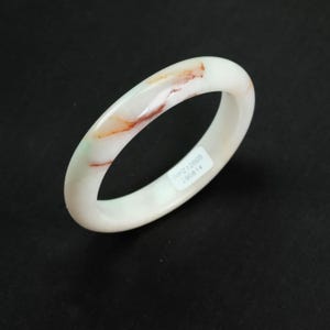 May include: A smooth, circular bangle bracelet crafted from off-white stone with natural reddish-brown and faint green veining throughout. A small rectangular white sticker is adhered to the inner surface, displaying the text WPZ12605 290814.