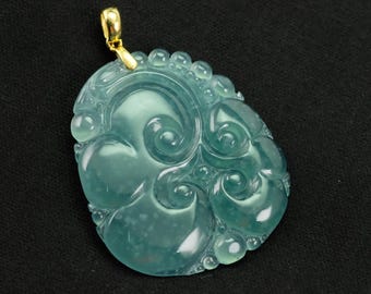 May include: A translucent, teal-colored jade pendant with intricate carved details. The pendant features a swirling design with rounded edges and a gold-colored bail for hanging. The jade has a polished, glossy finish.