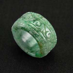 May include: A wide band ring carved from mottled light and dark green stone. The exterior surface features a continuous relief pattern of swirling scrolls and small circular indentations that wrap around the entire circumference of the ring.