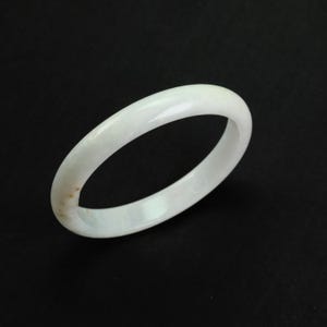 May include: A white jade bangle bracelet. The smooth, polished bracelet is a solid, circular band. The jade has a slightly translucent appearance with subtle variations in color. The background is a solid black.