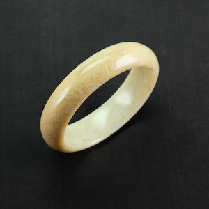 May include: A pale jade ring with a polished finish. The ring is a solid band, displaying a mix of pale yellow and cream hues. The ring's smooth surface highlights the natural variations in the stone's colour and texture.