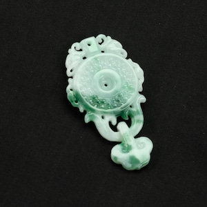 May include: A jade pendant with a light green and white color scheme. The pendant features a circular design with intricate carvings and a floral-like bottom element. The jade is polished and has a smooth surface, with a hole in the center of the circular design.