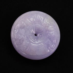 Puede incluir: Un colgante redondo de jade lavanda con intrincadas tallas. El colgante presenta un agujero central y patrones en relieve detallados, posiblemente representando diseños florales o simbólicos. La superficie pulida refleja la luz.