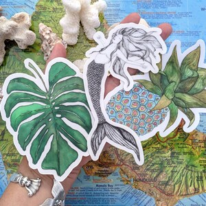 Op de afbeelding: Een set van vier ge&iuml;llustreerde stickers met een zeemeermin, een ananas, een monstera-blad en een ananas met een blauw en roze patroon. De stickers zijn op een kaart van Hawa&iuml;.