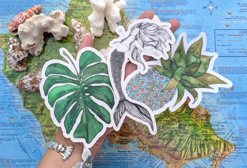 Op de afbeelding: Een set van vier ge&iuml;llustreerde stickers met een zeemeermin, een ananas, een monstera-blad en een ananas met een blauw en roze patroon. De stickers zijn op een kaart van Hawa&iuml;.