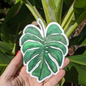 Op de afbeelding: Een groene monstera-bladsticker in aquarel. De sticker is op een witte achtergrond en heeft een licht getextureerd oppervlak.