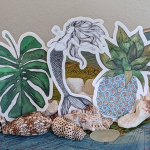 Op de afbeelding: Drie stickers met een zeemeermin, een ananas en een monstera-blad. De zeemeermin is in zwart-wit getekend en heeft lang, golvend haar. De ananas is getekend in een kleurrijke stijl met blauwe, roze en gele accenten. Het monstera-blad is getekend in tinten groen.