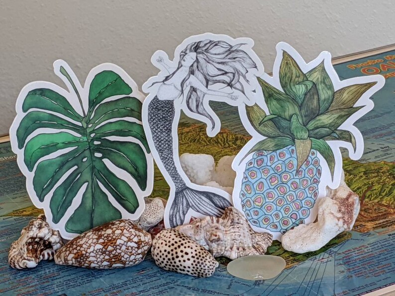Op de afbeelding: Drie stickers met een zeemeermin, een ananas en een monstera-blad. De zeemeermin is in zwart-wit getekend en heeft lang, golvend haar. De ananas is getekend in een kleurrijke stijl met blauwe, roze en gele accenten. Het monstera-blad is getekend in tinten groen.