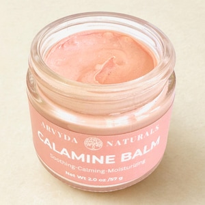 May include: A glass jar containing pink calamine balm. The jar has a pink label with the text "ARVYDA NATURALS CALAMINE BALM" and "Soothing-Calming-Moisturizing". The net weight is 57 g.
