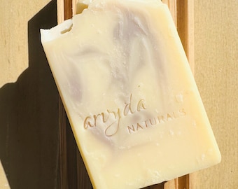 May include: A rectangular bar of soap with the text "aryida NATURALS" is placed on a wooden soap dish. The soap has a light yellow color with a marbled design. The soap dish is made of light-colored wood.