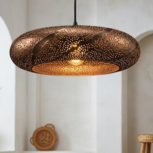 Op de afbeelding: Een ovale hanglamp in bronskleur met een gestructureerd, geperforeerd ontwerp. De lamp hangt aan een zwart snoer en straalt een warme gloed uit. Een decoratief element voor het interieur, ongeveer 40 cm in diameter.