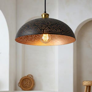 Puede incluir: Lámpara colgante de metal negro con diseño perforado e interior dorado. La lámpara cuelga de un cable negro con un accesorio dorado. La lámpara está en una habitación con una planta y un taburete tejido.