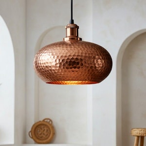 Pode incluir: Uma luminária pendente de cor cobre com textura martelada pende de um cabo preto. A luminária tem formato oval arredondado. O fundo apresenta arcos brancos e uma planta em vaso, sugerindo um tema de decoração de casa.
