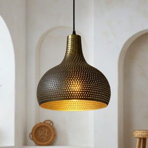 Puede incluir: Una lámpara colgante de color bronce con una pantalla perforada y un interior dorado. La luz cuelga de un cable negro contra una pared blanca con arcos. Una planta en maceta y objetos decorativos son visibles debajo.