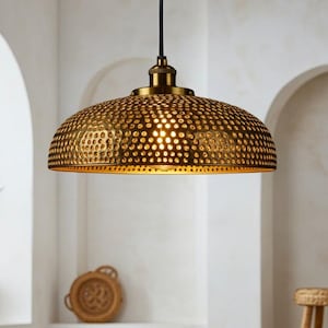 Könnte beinhalten: Eine goldfarbene, kuppelförmige Pendelleuchte mit strukturierter Oberfläche und kleinen Perforationen. Das Licht hängt an einem schwarzen Kabel vor einer weißen Wand mit gewölbten Nischen. Ein Blumentopf und ein geflochtener Tablett sind sichtbar.