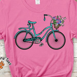 Puede incluir: Una bicicleta azul turquesa con una cesta de flores en el manillar. La bicicleta tiene un fondo rosa.
