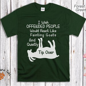 Może przedstawiać: Leśnozielona koszulka z białym napisem: "I Wish Offended People Would React Like Fainting Goats And Quietly Tip Over." Poniżej tekstu znajduje się biała grafika przedstawiająca kozę. Koszulka jest na drewnianym tle.