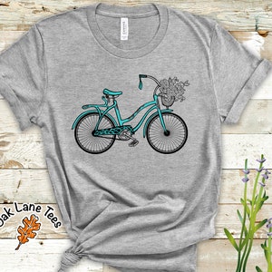 May include: A teal blue bicycle with a basket full of flowers on a gray t-shirt. The t-shirt has the text "Oak Lane Tees" with a brown leaf design.