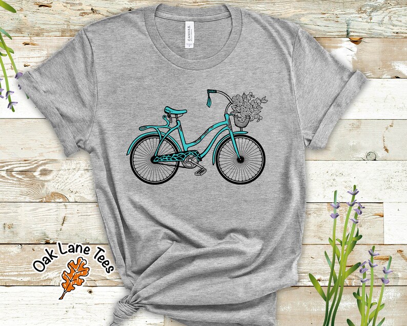 May include: A teal blue bicycle with a basket full of flowers on a gray t-shirt. The t-shirt has the text "Oak Lane Tees" with a brown leaf design.